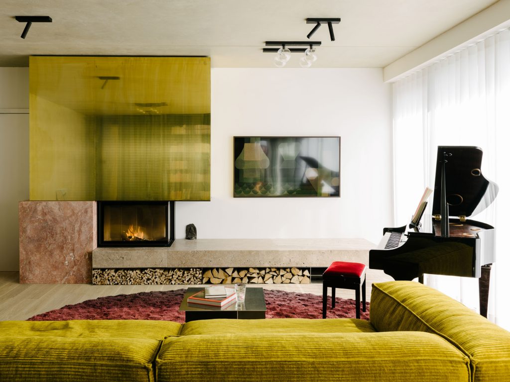 The Living Room occupies the space between an existing wall and the green box. The hearth is made from travertine Sierra ebru stone, red travertine, brass, and thin plates of stainless steel. Te design strategy is to create relationship between boxes. Photo ©Robert Rieger, Courtesy of Ester Bruzkus Architekten.