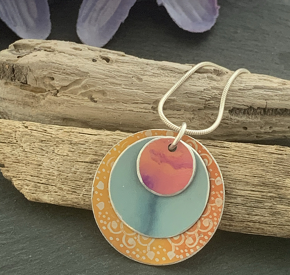 Hand Painted Aluminium Pendant, Teal and Orange Mandala by the Cheshire Jewellery Company
 This pendant is made with hand dyed/painted aluminium and sterling silver chain. The pendant is made with 3 layered discs. Each layer has been hand painted. The larger lower disc is orange with a mandala print. The middle layer is duck egg blue/grey and the top layer is orange with flecks of dark pink. All 3 shapes have a gentle curve to them and sit one top of each other. The width of the lower larger disc is approximately 2.5 cm.